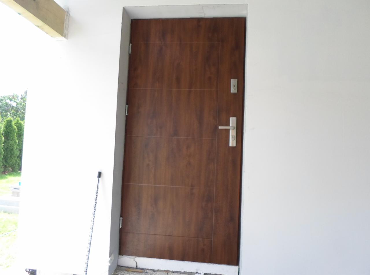 Zamontowane brązowe drzwi wejściowe w białej ścianie budynku. Widoczne detale zamka i klamki. Częściowo widoczny fragment elewacji i zieleń w tle.