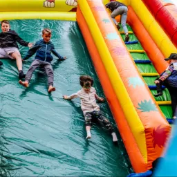 Dzieci zjeżdżają z dmuchanej zjeżdżalni; jeden chłopiec ma pomalowaną twarz na zielono.