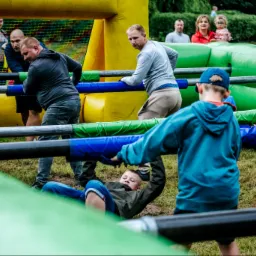 Grupa osób, w tym dzieci, bierze udział w grze w piłkarzyki stołowe na dmuchanym boisku podczas imprezy plenerowej, z widocznymi elementami dmuchanych konstrukcji w kolorach żółtym, zielonym...