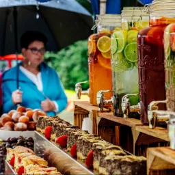 Stół z napojami w dużych słojach z kranikami, ciastami i owocami, przygotowany na zewnątrz, z osobami w tle pod parasolem.