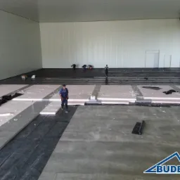 Montaż izolacji podłoża w hali przemysłowej. Widoczne rozłożone płyty styropianowe, czarna folia i pracownicy. Budex S.C. w prawym dolnym rogu.