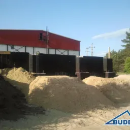 Wykop pod fundamenty z zabezpieczeniem szalunkowym i hałdami piasku na tle hali przemysłowej w słoneczny dzień. Widoczne zbrojenia.