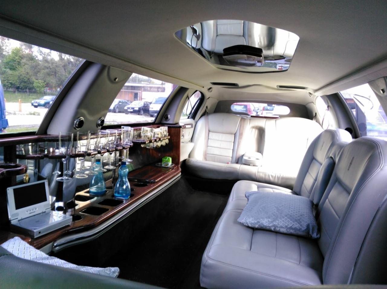 Wnętrze luksusowej limuzyny z barem, szklankami, napojami i skórzanymi siedzeniami. Widok na okno i inne pojazdy na zewnątrz. Komfort i elegancja w podróży.