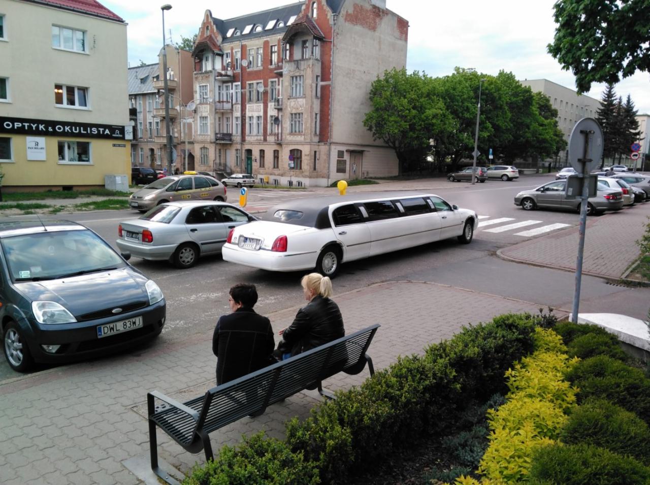 Biała limuzyna skręca na skrzyżowaniu w Olsztynie, widoczna zza dwóch kobiet siedzących na ławce.