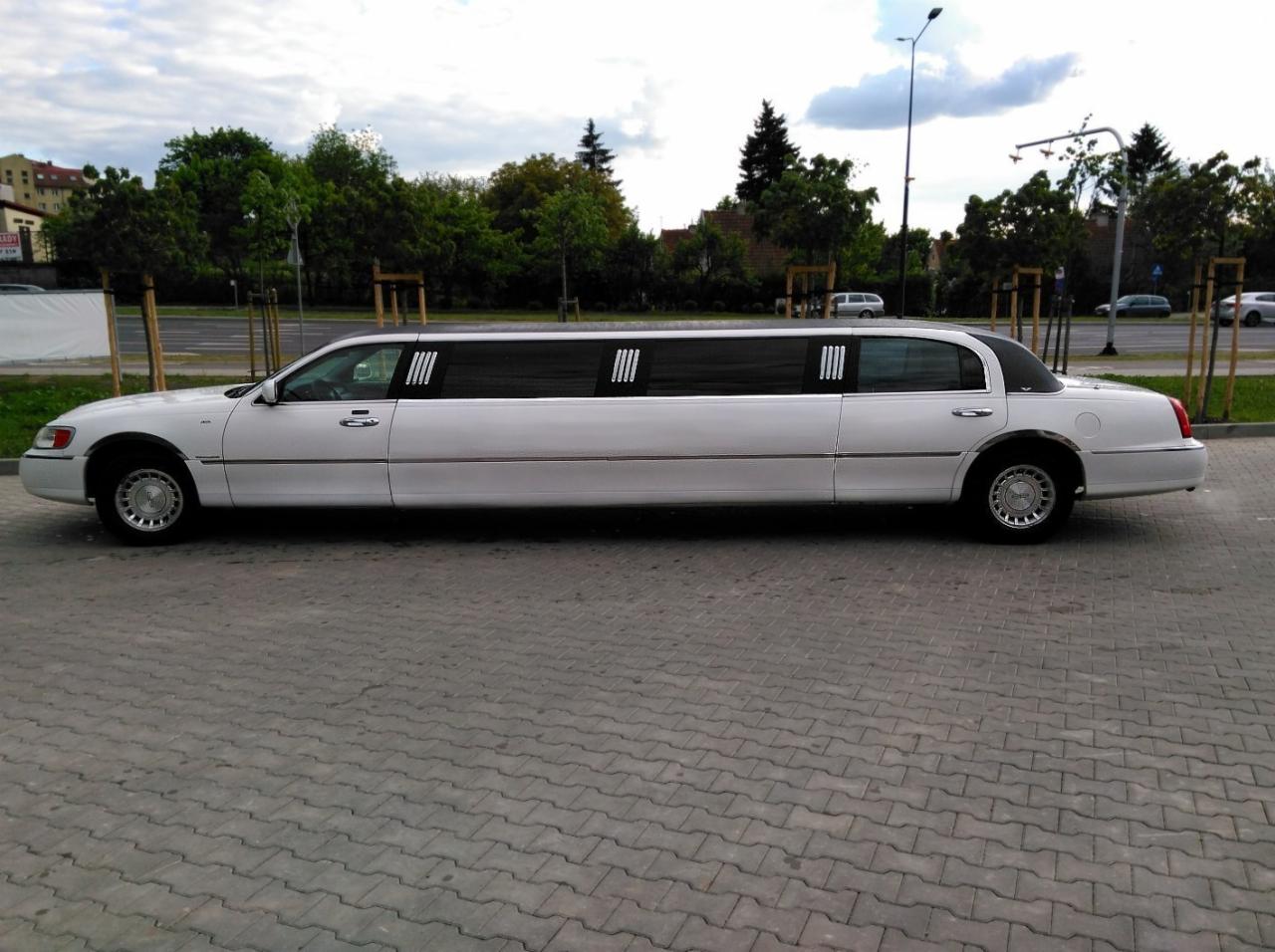 Elegancka, biała limuzyna Lincoln Town Car z przyciemnianymi szybami, zaparkowana na brukowanej powierzchni. Widok z boku, tło stanowią drzewa i budynki.