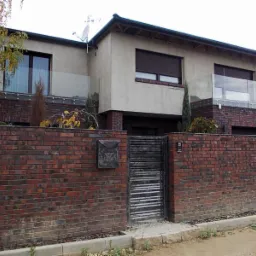 Dwukondygnacyjny dom z ceglanym ogrodzeniem i ciemnymi roletami w oknach. Balkony z szklanymi balustradami. Skrzynka na listy na ogrodzeniu.