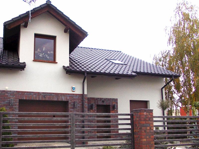 Dwukondygnacyjny dom z ciemną dachówką, ceglaną fasadą garażu i metalowym ogrodzeniem. Widoczne okna z brązowymi ramami i system rynnowy.