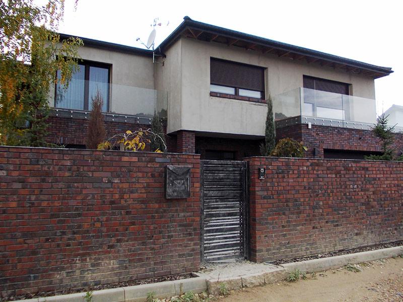 Dwukondygnacyjny dom z ceglanym ogrodzeniem i ciemnymi roletami w oknach. Balkony z szklanymi balustradami. Skrzynka na listy na ogrodzeniu.