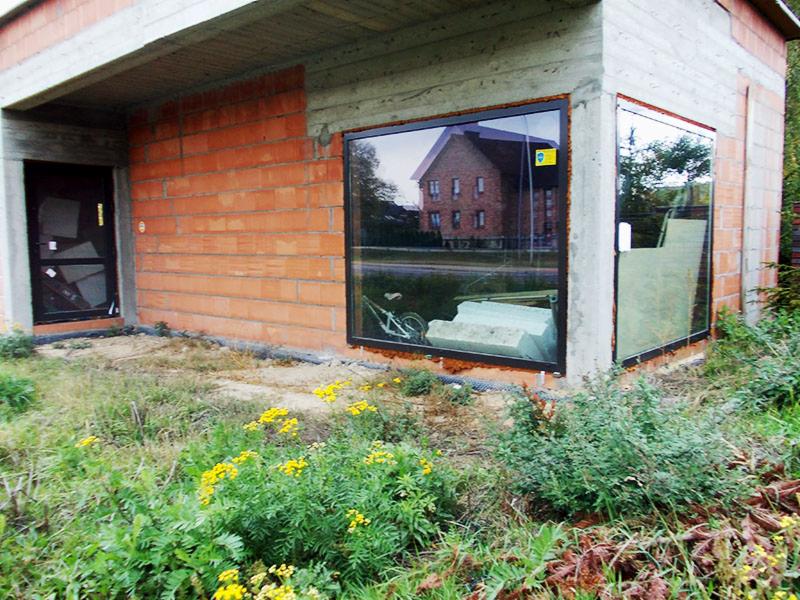Surowy budynek z zamontowanymi oknami z czarnymi ramami. W oknach odbija się sąsiedni dom. Na zewnątrz widoczne żółte kwiaty i trawa.