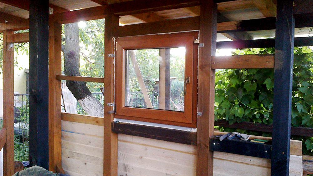 Montaż okna drewnianego w konstrukcji drewnianej altany, widoczne elementy mocujące i fragment ogrodu w tle. Świeże, jasne drewno kontrastuje z ciemnymi elementami.