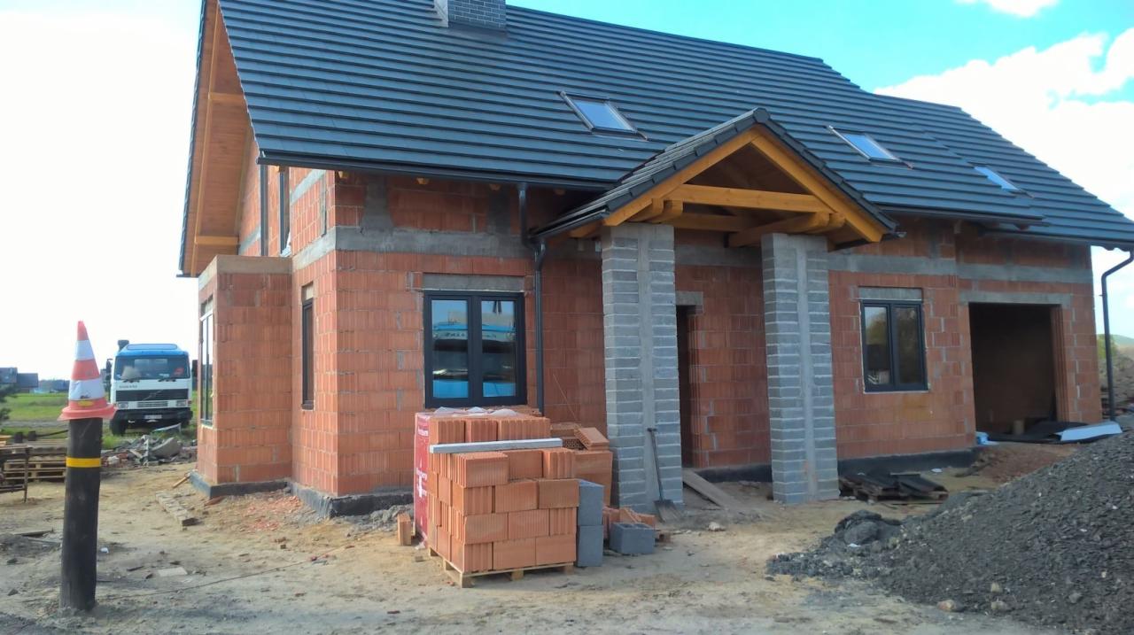 Surowy, dwukondygnacyjny dom z cegły w trakcie budowy. Widoczne okna, dach z dachówką i sterta cegieł. Na pierwszym planie słupek z pachołkiem.