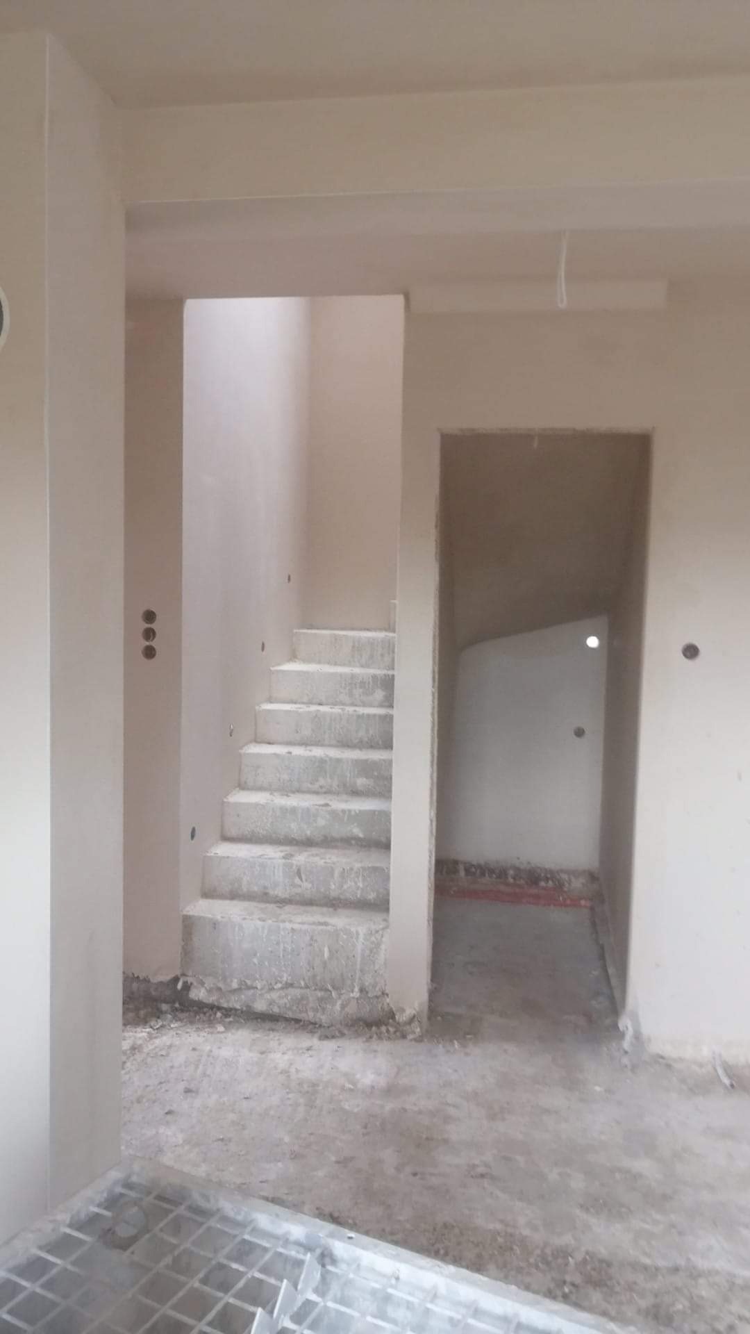Surowe schody betonowe w nowo wykończonym wnętrzu, ściany pokryte świeżym tynkiem, widoczne otwory na oświetlenie, podłoga częściowo zakryta metalową kratką.