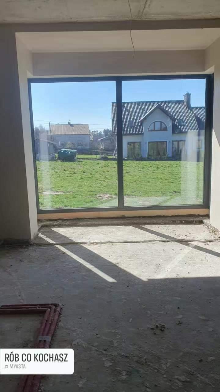 Wnętrze surowego domu w trakcie budowy z widokiem przez duże okno na ogród i domy w tle, rury instalacyjne leżą na podłodze.