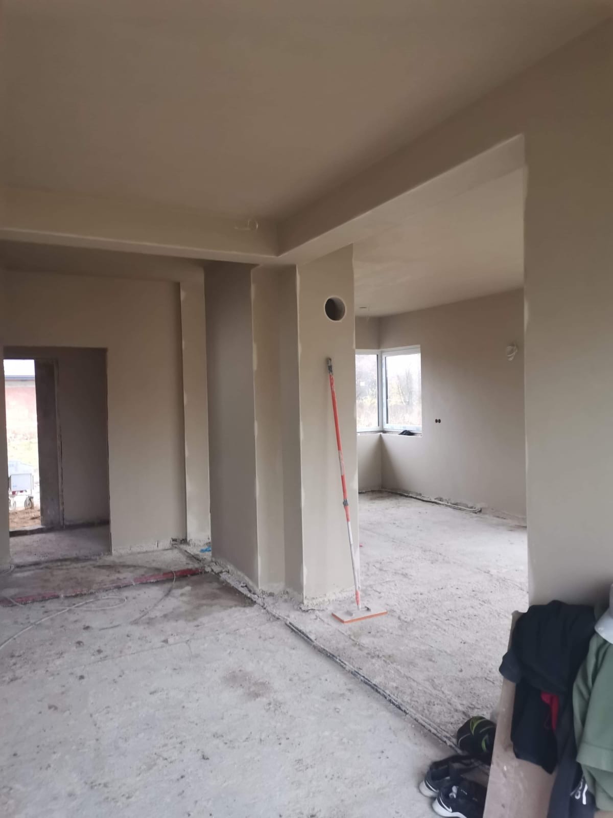 Wnętrze w stanie deweloperskim z nałożonymi jasnymi tynkami, widoczny wałek do rozprowadzania masy szpachlowej oparty o ścianę, przewody elektryczne ułożone na podłodze.