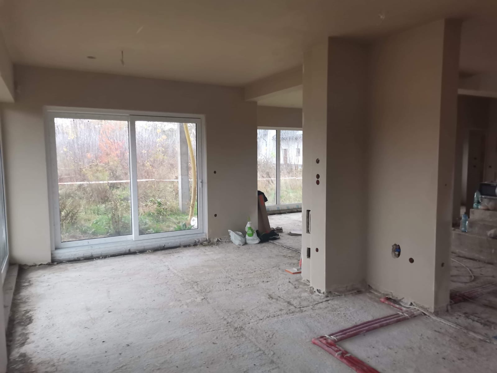 Wnętrze domu w stanie deweloperskim po tynkowaniu maszynowym, widoczne okna przesuwne, instalacja ogrzewania podłogowego i otwory pod gniazdka elektryczne.