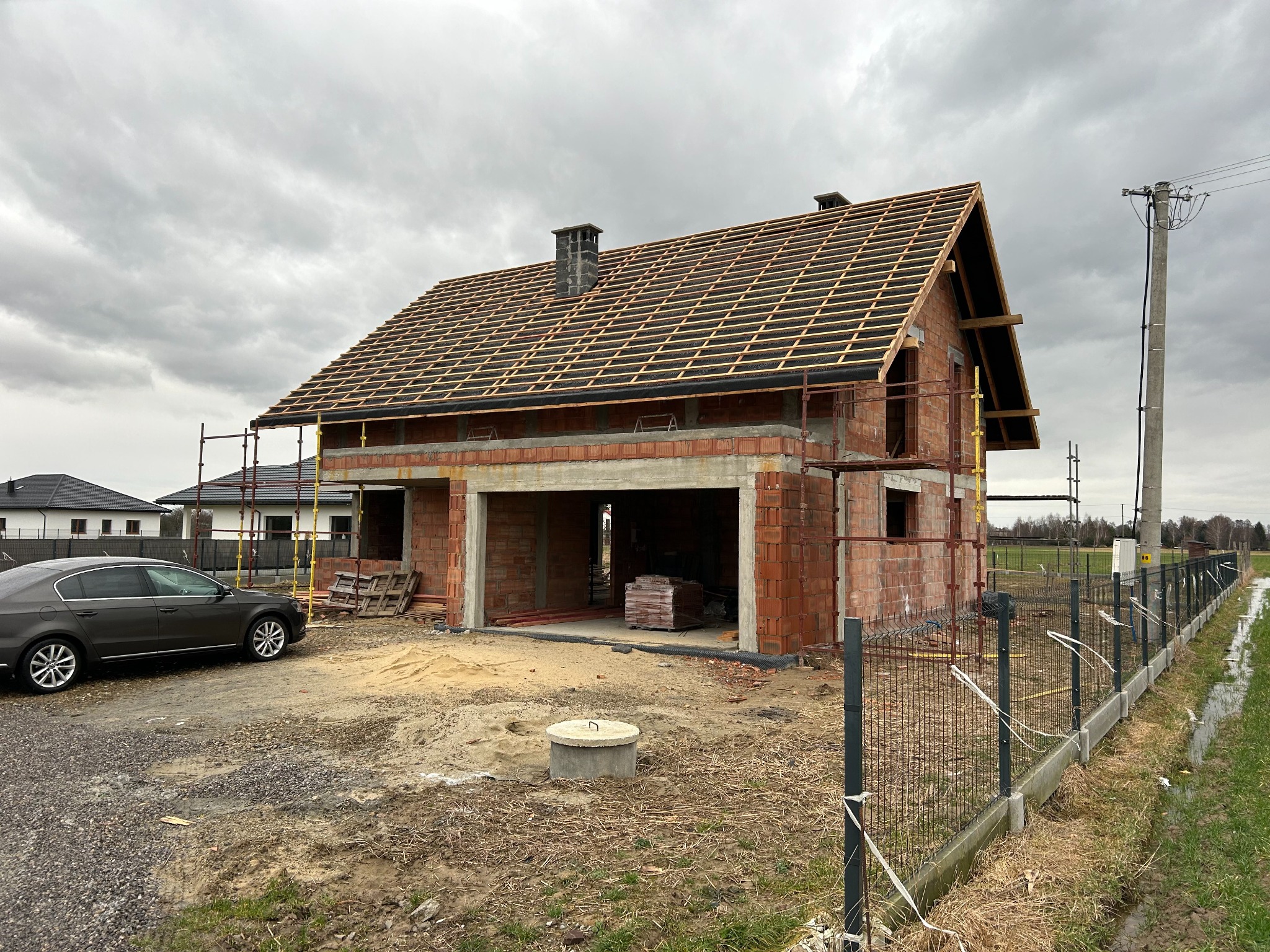 Surowy stan domu jednorodzinnego z czerwonej cegły, z widoczną konstrukcją dachu z łat, pod pochmurnym niebem, samochód osobowy zaparkowany obok placu budowy.