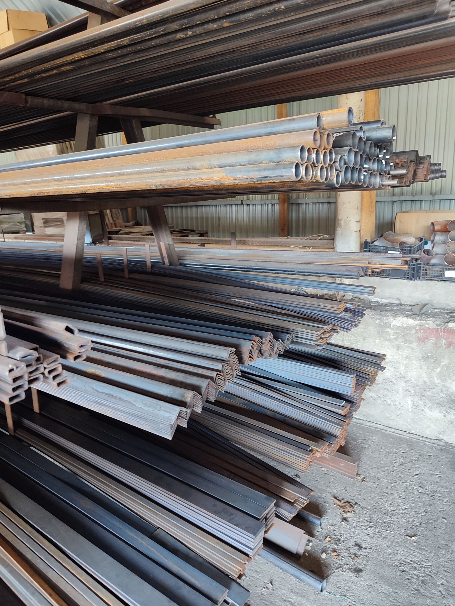 Różnorodne profile stalowe (rury okrągłe, płaskowniki, kątowniki) składowane na regale w magazynie, widoczne ślady korozji na niektórych elementach.