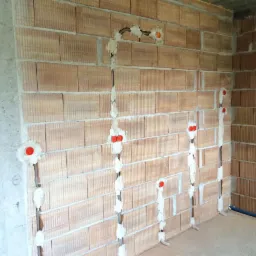 Ściana z pustaków ceramicznych z bruzdami wypełnionymi gipsem, w których poprowadzone są przewody elektryczne zakończone pomarańczowymi puszkami instalacyjnymi.