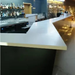 Długi, biały blat baru z czarną podstawą w lokalu gastronomicznym, widoczne butelki alkoholi i szklanki, oświetlenie LED pod sufitem.