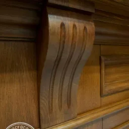 Detal drewnianej boazerii z dekoracyjnym wspornikiem z trzema wyżłobieniami, widoczny fragment logotypu firmy w lewym dolnym rogu.