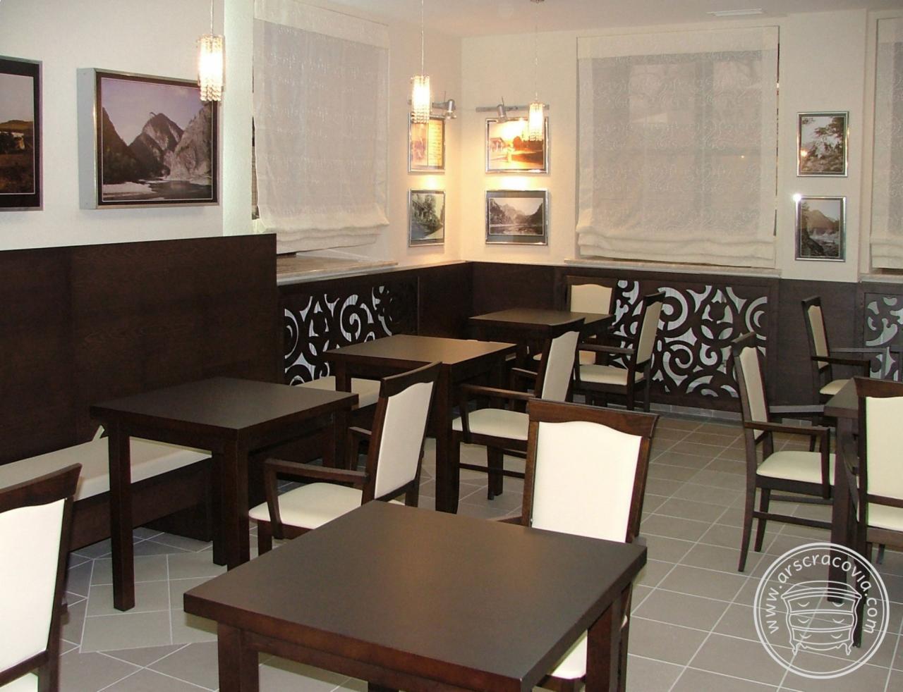 Wnętrze kawiarni z ciemnymi drewnianymi stolikami i krzesłami z jasnymi obiciami, dekoracyjna ciemna boazeria z wycinanym wzorem, jasne rolety rzymskie w oknach i obrazy na ścianach.