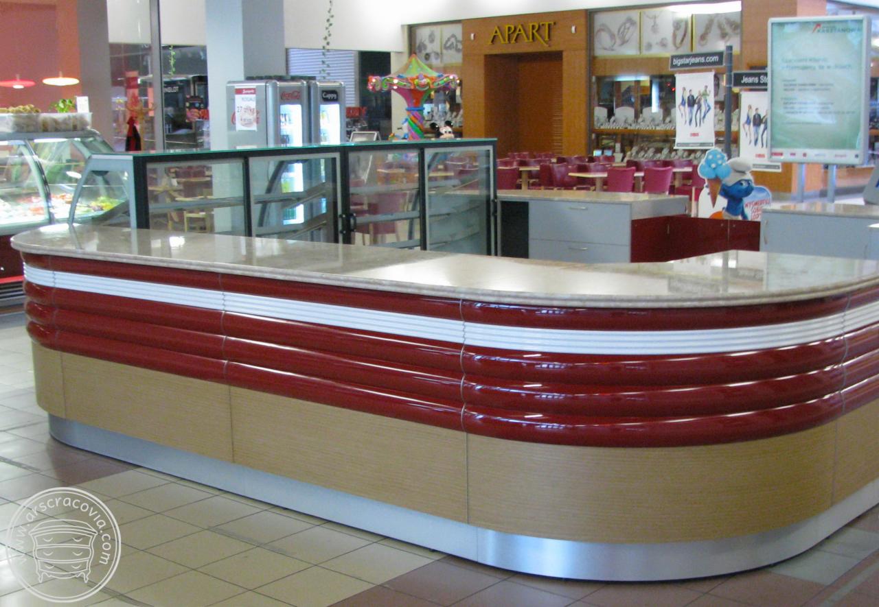 Zakrzywiona lada recepcyjna z marmurowym blatem, czerwonymi i białymi detalami oraz drewnopodobną podstawą, w tle witryny chłodnicze i wnętrze centrum handlowego.