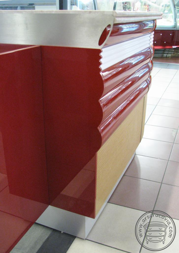 Czerwona lada barowa z falistymi zdobieniami i metalowym blatem, widoczna z boku, ustawiona na podłodze wyłożonej kafelkami.