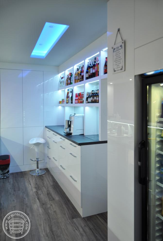 Biała zabudowa baru z podświetlanymi półkami wypełnionymi butelkami alkoholi, ekspres do kawy na blacie, hokery i chłodziarka na wino, podświetlenie LED w suficie.
