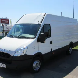 Biały samochód dostawczy Iveco Daily z wysokim dachem, zaparkowany na brukowanym parkingu w słoneczny dzień, widoczny znak 'Wyjazd' w tle.