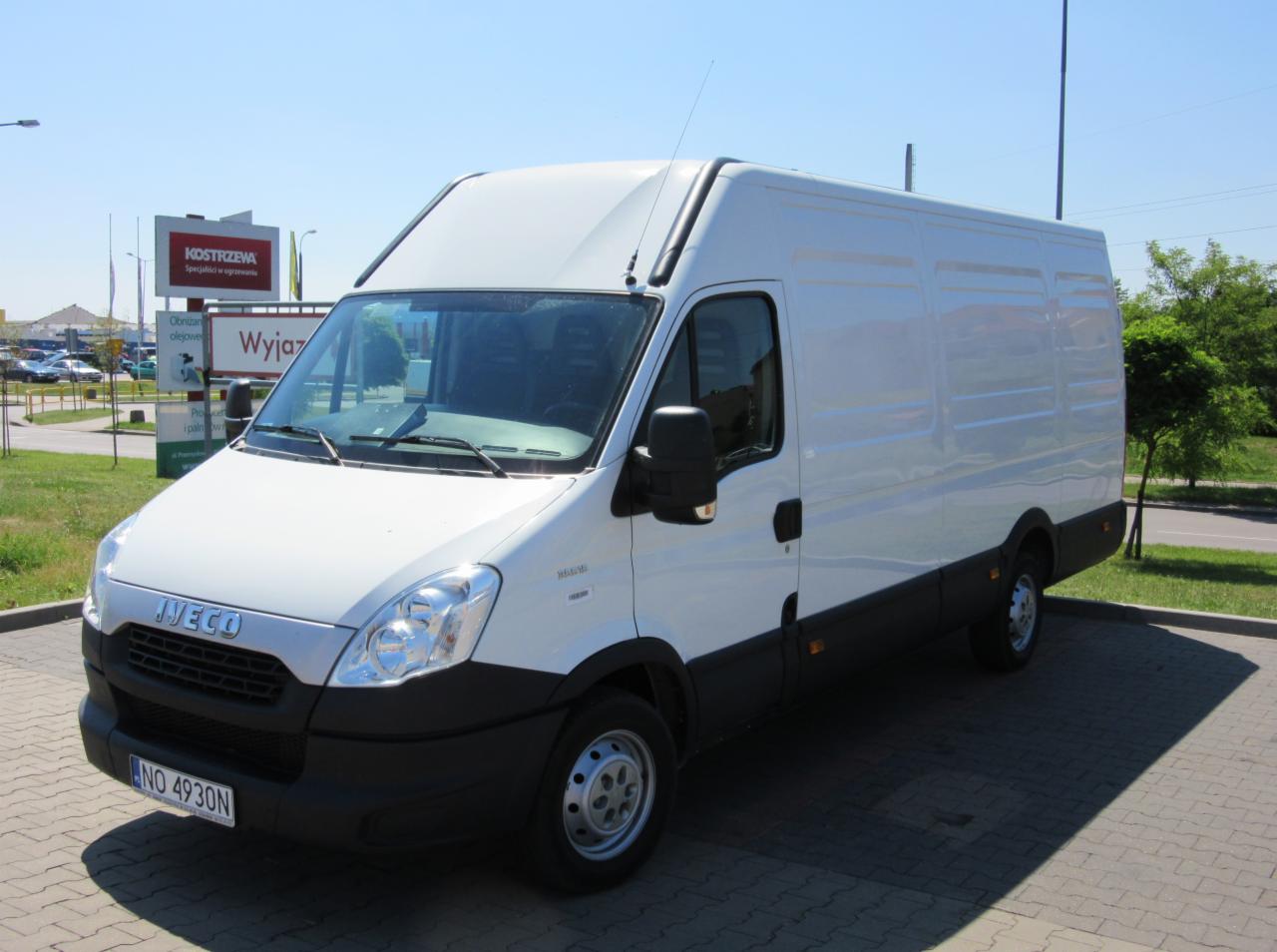 Biały samochód dostawczy Iveco Daily z wysokim dachem, zaparkowany na brukowanym parkingu w słoneczny dzień, widoczny znak 'Wyjazd' w tle.