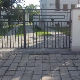 Nowoczesna, dwuskrzydłowa brama wjazdowa z metalowymi prętami i dekoracyjnymi prostokątami, osadzona na białych, murowanych słupkach z domofonem. Podjazd wyłożony kostką brukową, w tle budynek...