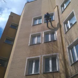 Pracownik na linie zabezpieczającej myje elewację budynku z cegły, widoczne zacieki wody na ścianie.