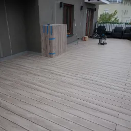 Jasnoszary taras na dachu budynku wielorodzinnego, z widocznym fragmentem elewacji, meblami ogrodowymi i sprzętem budowlanym, częściowo zabezpieczony niebieską taśmą.