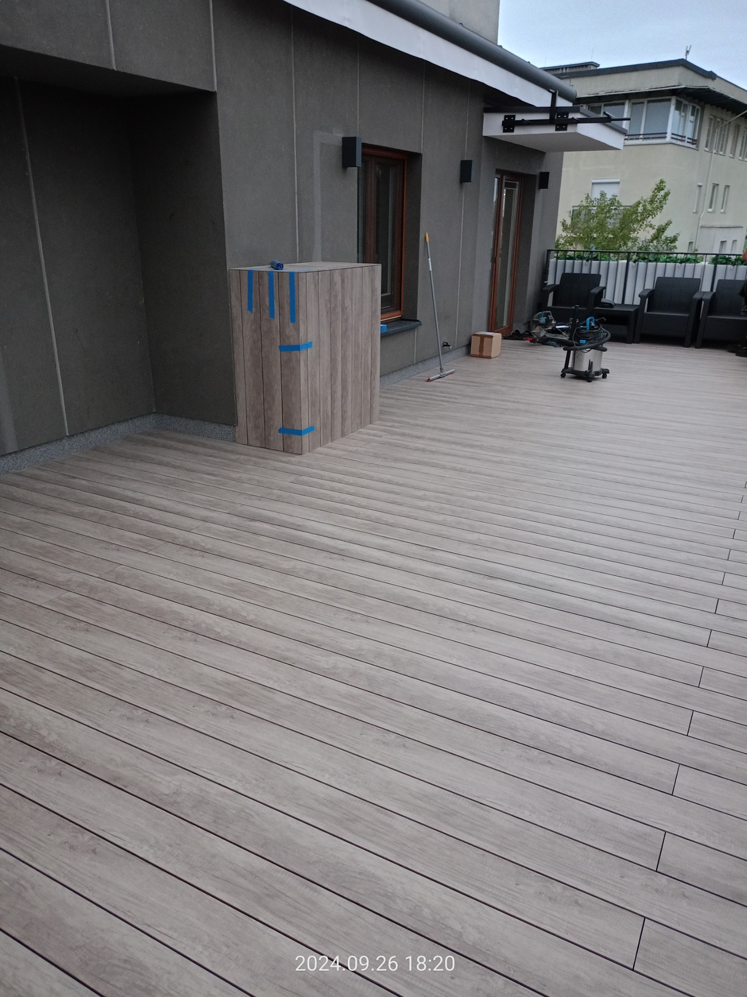 Jasnoszary taras na dachu budynku wielorodzinnego, z widocznym fragmentem elewacji, meblami ogrodowymi i sprzętem budowlanym, częściowo zabezpieczony niebieską taśmą.