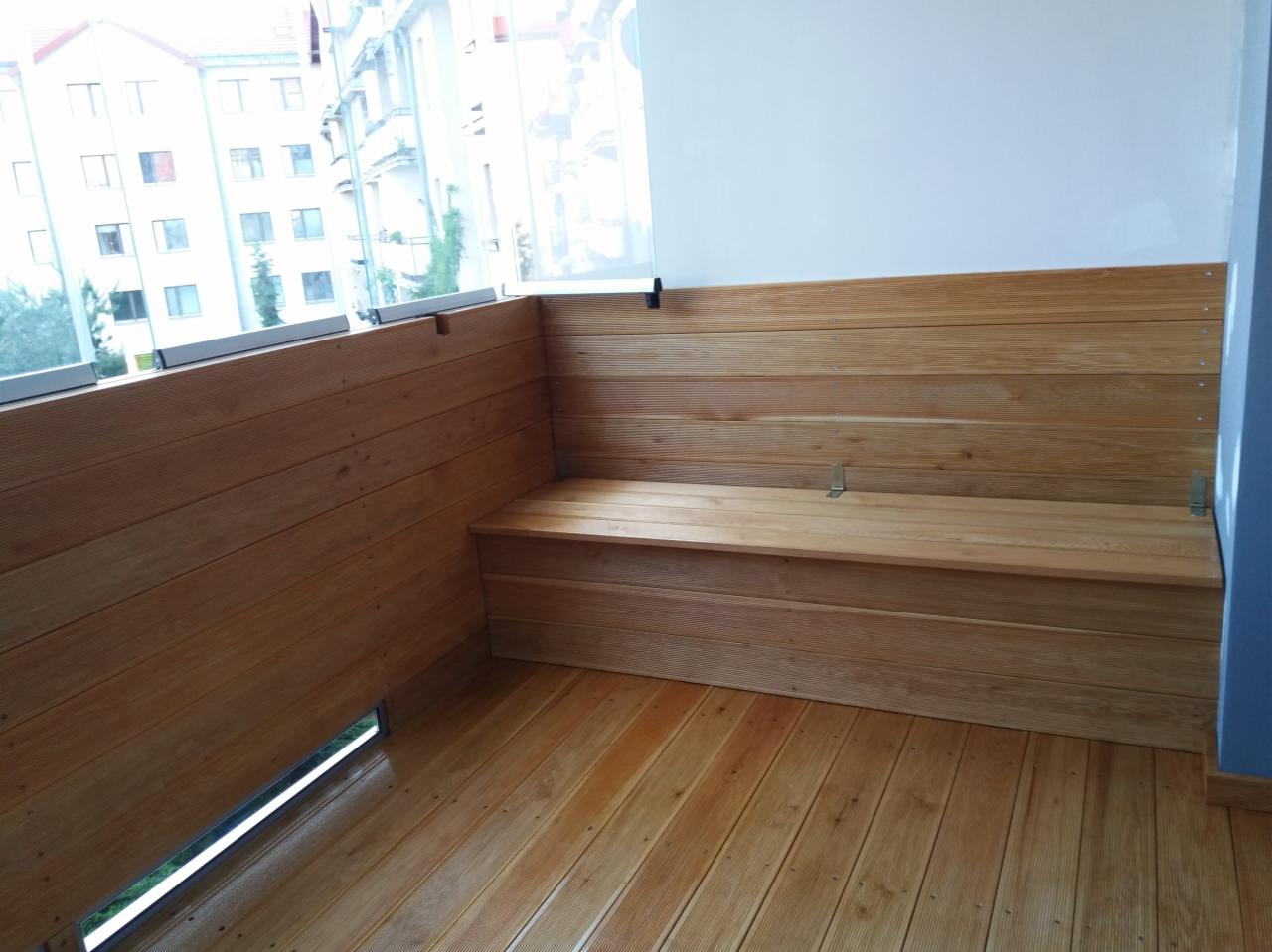 Zabudowa drewniana balkonu z siedziskiem i drewnianą podłogą, widok na budynek przez szklaną balustradę.