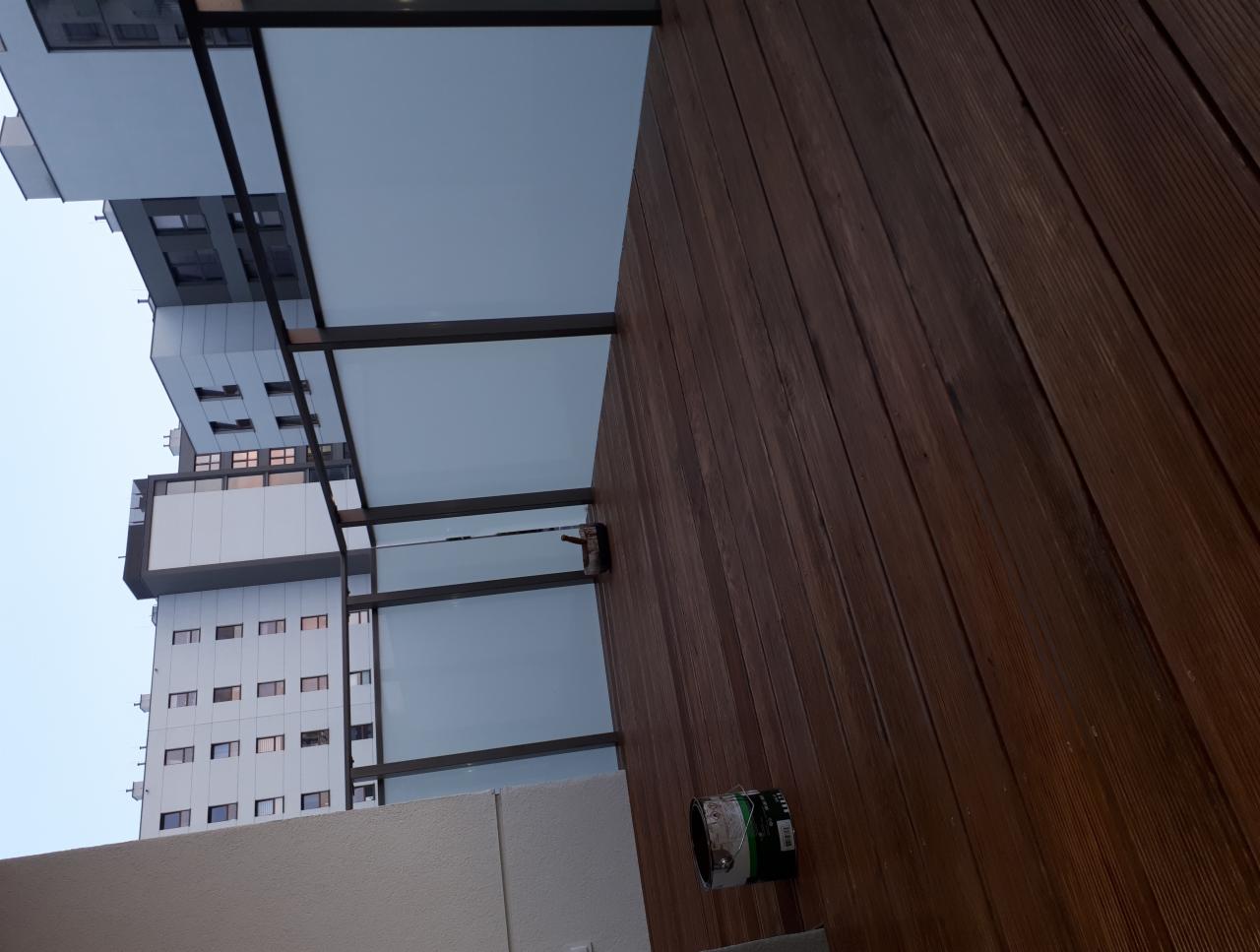 Zadaszony taras drewniany z widokiem na wysoki budynek mieszkalny, puszka farby leżąca na podłodze tarasu.