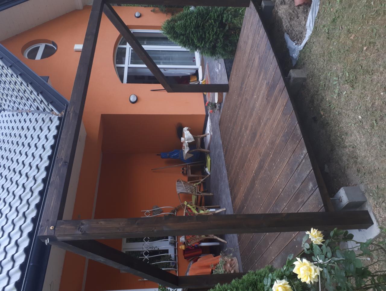 Ciemnobrązowa konstrukcja pergoli z drewnianym tarasem, widok na pomarańczowy dom z zaokrąglonym oknem i krzesłami na zewnątrz, w tle trawnik i roślinność.