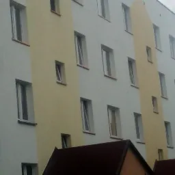 Wielopiętrowy budynek z oknami, elewacja w jasnych odcieniach z żółtym akcentem. Widoczne fragmenty dachu mniejszego budynku na pierwszym planie. Ujęcie z dołu.