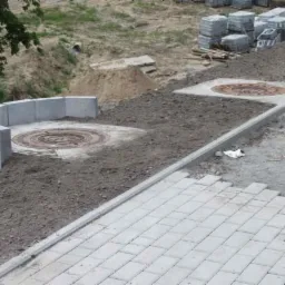 Etap układania kostki brukowej z obrzeżami i studzienkami kanalizacyjnymi. Częściowo ułożona nawierzchnia z kostki, stosy kostki w tle, teren przygotowany.