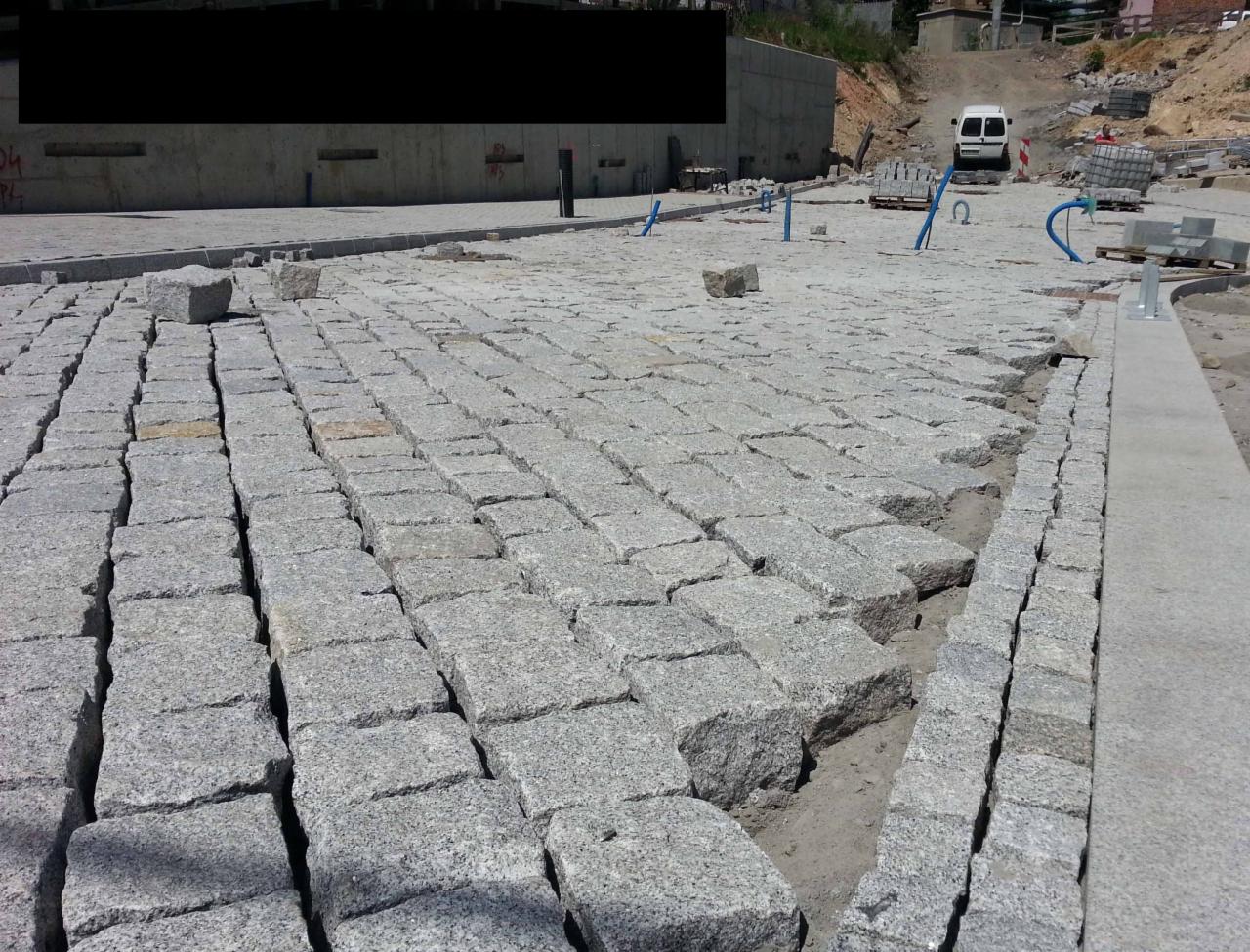 Układanie kostki granitowej na placu, widoczne przestrzenie między kostkami, przygotowane podłoże. W tle betonowy mur i biały bus.