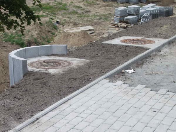 Etap układania kostki brukowej z obrzeżami i studzienkami kanalizacyjnymi. Częściowo ułożona nawierzchnia z kostki, stosy kostki w tle, teren przygotowany.