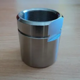 Cylindryczny, metalowy element z gwintem wewnętrznym i wyfrezowaną krawędzią, leżący na drewnianej powierzchni.
