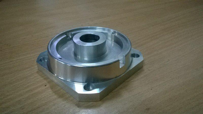 Precyzyjnie obrobiony, srebrny element metalowy z otworem centralnym i czterema otworami montażowymi na podstawie, leżący na drewnianej powierzchni.