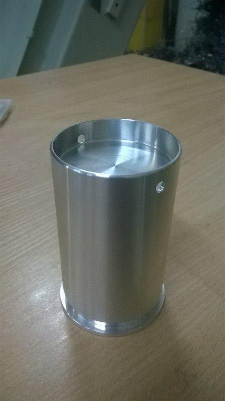 Cylindryczny, metalowy element z dwoma otworami, obrobiony z widocznymi śladami frezowania na górnej powierzchni, stojący na drewnianym blacie.