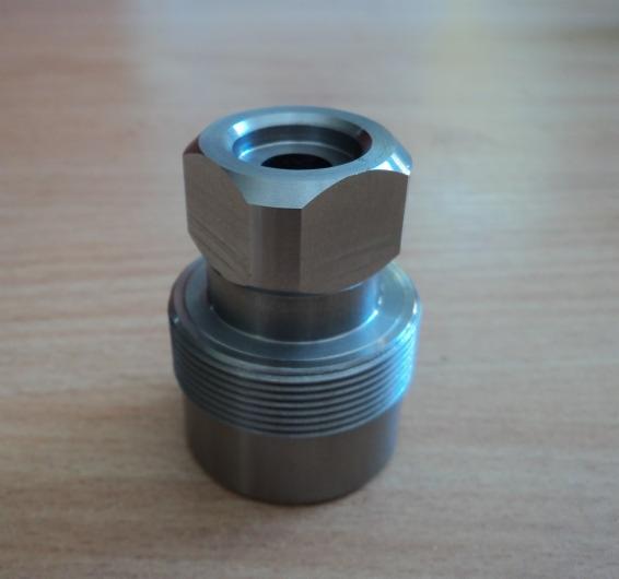 Precyzyjnie obrobiony, metalowy element z gwintowanymi żłobieniami na dolnej części i sześciokątną nakrętką na górze, leżący na drewnianym blacie.