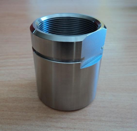 Cylindryczny, metalowy element z gwintem wewnętrznym i wyfrezowaną krawędzią, leżący na drewnianej powierzchni.