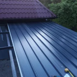 Montaż paneli warstwowych na konstrukcji stalowej dachu, widoczna wkrętarka i miara, z częściowo widocznym fragmentem dachu pokrytego bordową blachodachówką.