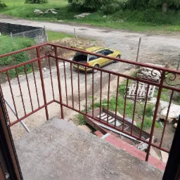 Czerwona metalowa balustrada balkonowa z ozdobnymi elementami, widok z balkonu na podwórko z zaparkowanym żółtym samochodem z otwartym bagażnikiem, w tle zieleń i droga.