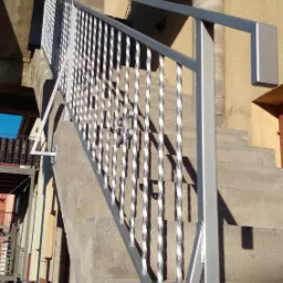 Zewnętrzne schody betonowe z nowoczesną balustradą ze stali nierdzewnej o spiralnych wypełnieniach pionowych, przytwierdzoną do boku schod&oacute;w.