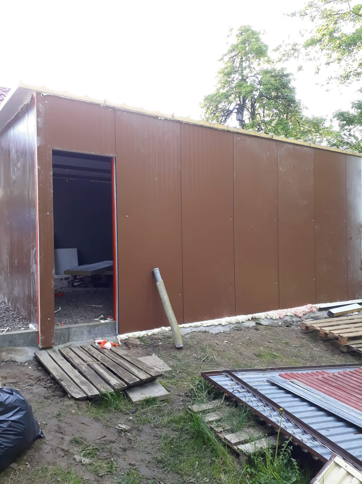 Budowa hali z brązowych płyt warstwowych, widoczne płyty ułożone na ziemi, otwór drzwiowy bez drzwi i fragment dachu, otoczenie budowy w trakcie realizacji.